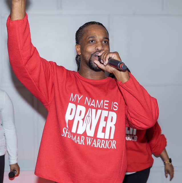 A worshipper in a red My Name Is Prayer Shamar Warrior long-sleeve shirt lifting his hand