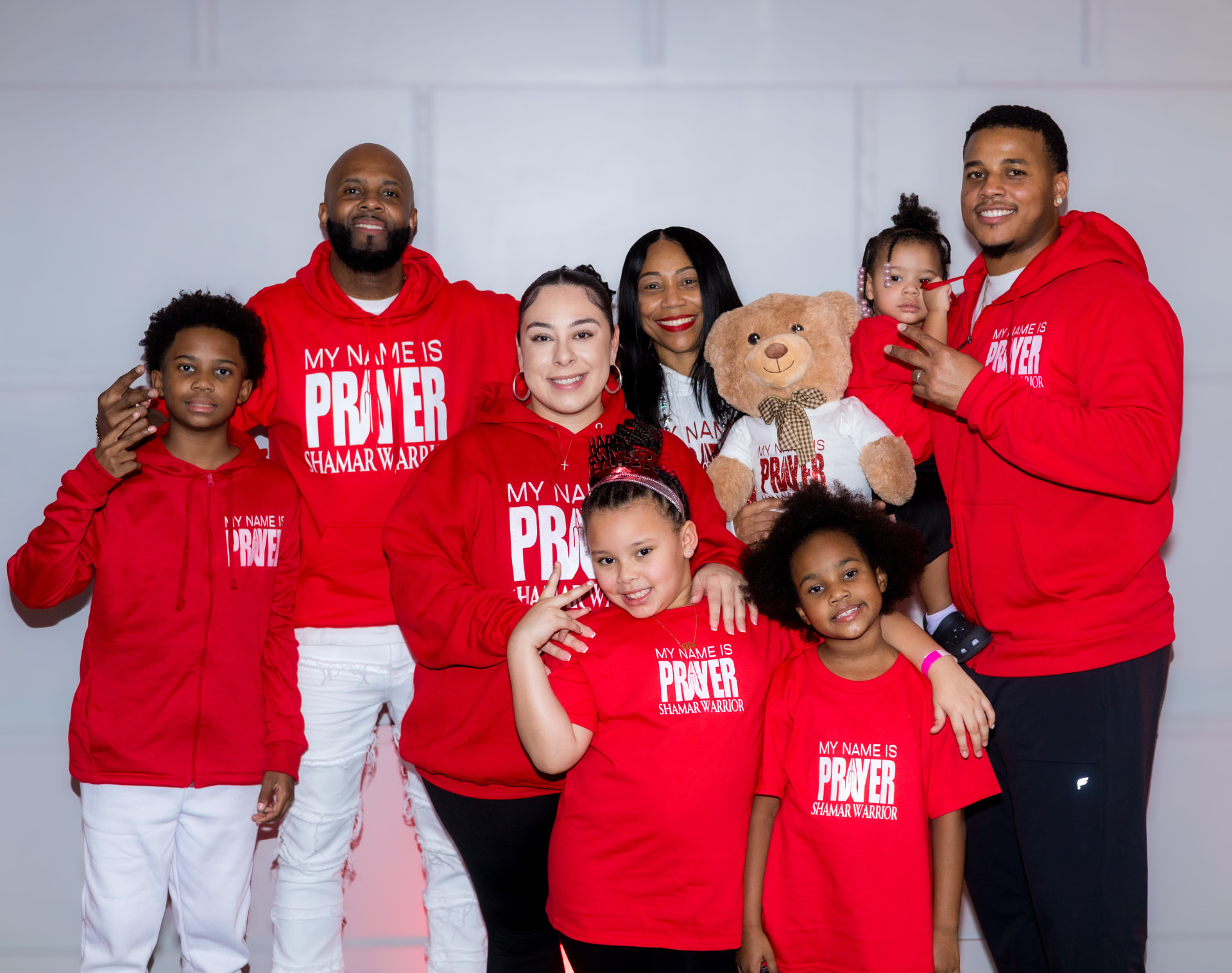 A family wearing matching red My Name Is Prayer hoodies and tees with the Prayer Comfort Bear