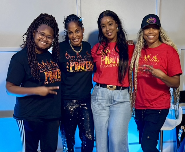 Four women wearing Light The Flame My Name Is Prayer tees in black and red