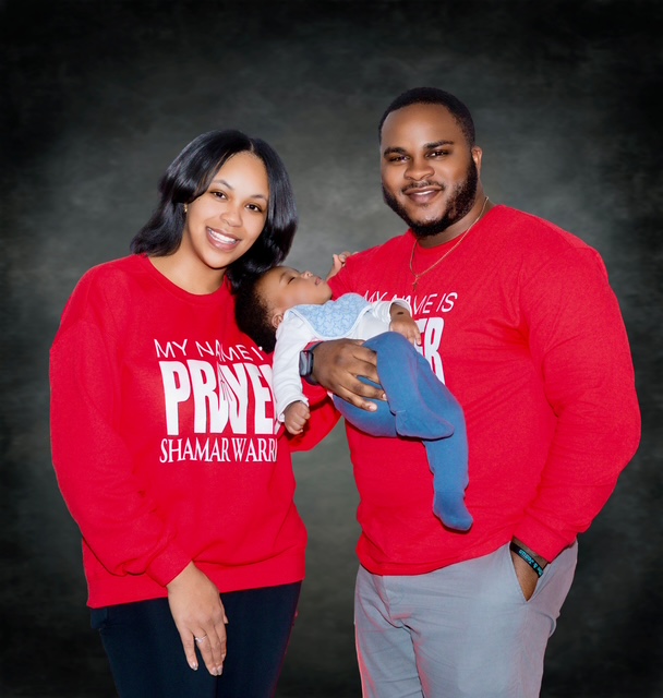 Parents holding their baby while wearing matching red My Name Is Prayer long-sleeve shirts
